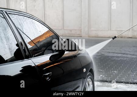 Lavare un'auto nera di lusso con un autolavaggio senza contatto. Pulizia dei dettagli dell'auto. Lavaggio auto berlina con schiuma self-service e acqua ad alta pressione. Pulizia e ordine in ambiente urbano Foto Stock