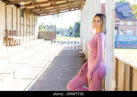 Una giovane donna vestita in forma rosa si appoggia contro una colonna in un ambiente urbano di palestra, il suo sguardo è fissato con sicurezza in lontananza. Athletic Poise: Donna sicura di sé in outfit fitness presso Urban Gym. Foto di alta qualità Foto Stock