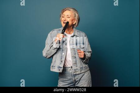 Allegra donna anziana con capelli grigi balla e canta in uno studio. È vestita con un abbigliamento casual in denim e ha una spazzola per capelli. La sua giocosa e la sua cura Foto Stock