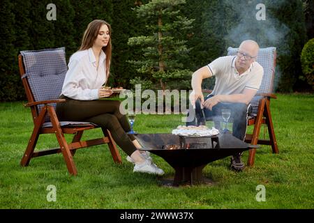 Coppia felice barbeuing all'aperto. Uomo che gira le salsicce sul grill. Foto Stock