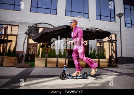 Donna matura vestita con stile con un pantalone rosa brillante di pantaloni e una giacca girano per la città su uno scooter, un frien attivo, sano ed ecologico Foto Stock