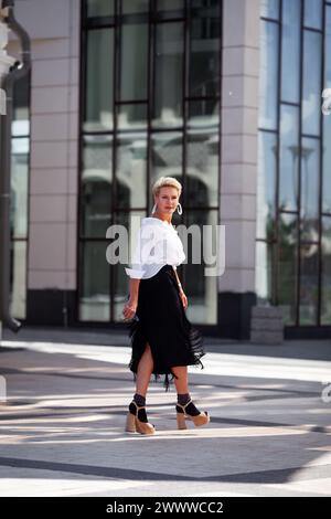 Elegante fashionista di mezza età, vestito con un abbigliamento alla moda gonna nera e una camicetta bianca, passeggia per strada, guardando indietro con eleganza. Bella matur Foto Stock