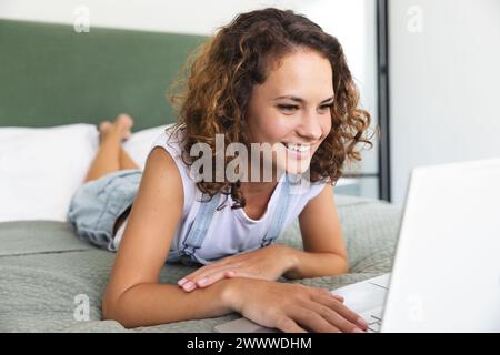 Donna caucasica interagisce con un portatile, sdraiata sul letto di casa Foto Stock