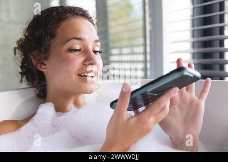 Donna caucasica si rilassa in un bagno di bolle a casa, smartphone in mano Foto Stock