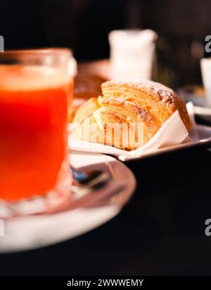 Un croissant fresco seduto su un piatto con un tovagliolo sotto e una tazza di succo d'arancia con un cucchiaio accanto sul lato sinistro. Foto Stock