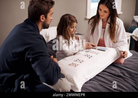 Genitori, ragazza e bambino con carte da gioco in camera da letto per legare, imparare e rilassarsi con amore in casa. Padre, madre e figlia con i giochi Foto Stock