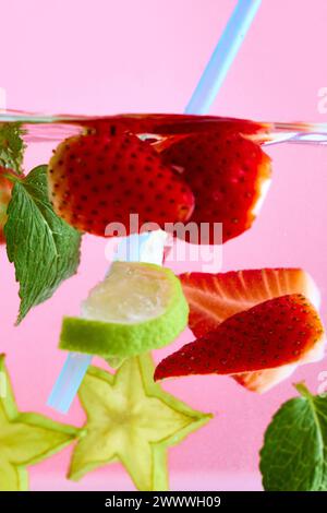 Fragole a fette e menta verde che galleggiano nell'acqua con paglia azzurra su sfondo rosa. Consistenza di una bevanda fresca dolce estiva. Foto Stock