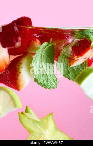 Fragole a fette, lime, limone, frutta stellata e menta verde che galleggiano nell'acqua con paglia azzurra su sfondo rosa. Consistenza del dolce rinfrescante Foto Stock