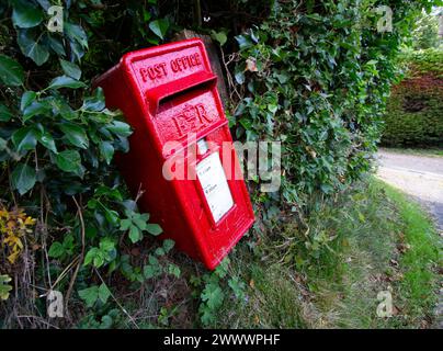 Sul lato di una strada di campagna, una cassetta delle lettere delle poste britanniche di colore rosso brillante si inclina in un angolo eccentrico Foto Stock