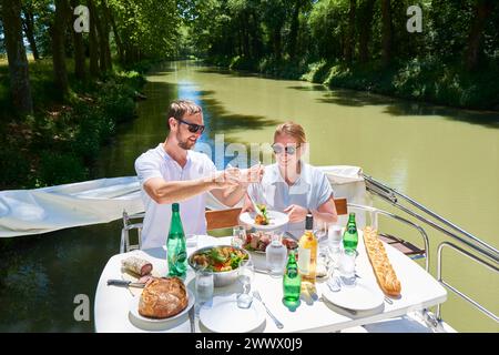 Coppia su una chiatta a noleggio sul Canal du Midi. Pasto a bordo dopo l'ormeggio lungo le rive delimitate da platani. Foto Stock