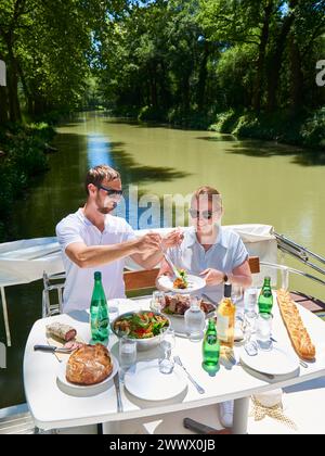 Coppia su una chiatta a noleggio sul Canal du Midi. Pasto a bordo dopo l'ormeggio lungo le rive delimitate da platani. Foto Stock