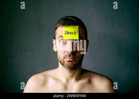 Un uomo confuso e scioccato con una nota adesiva gialla sulla fronte su sfondo grigio Foto Stock