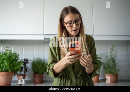Sorpresa donna di mezza età scioccata guardando lo schermo dello smartphone riceve un messaggio a casa. Foto Stock