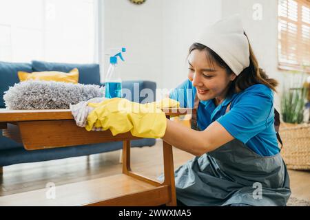 Con un sorriso, una donna diligente con guanti gialli pulisce e pulisce il tavolo del salotto. Lei Foto Stock