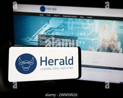 Persona che detiene lo smartphone con il logo della società britannica Herald Investment Trust plc davanti al sito Web. Mettere a fuoco il display del telefono. Foto Stock