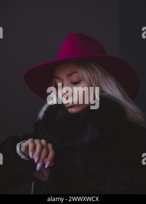 Giovane donna bionda contemplativa con cappello rosso e collo in pelliccia con illuminazione viola Foto Stock