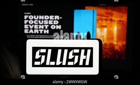 Persona che detiene un cellulare con il logo dell'evento finlandese annuale di startup e tecnologia Slush di fronte alla pagina web dell'azienda. Mettere a fuoco il display del telefono. Foto Stock