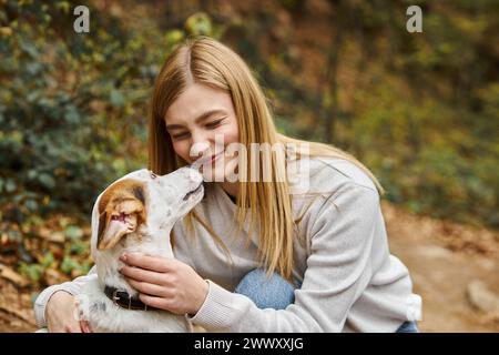 Donna emotiva che bacia e abbraccia il suo carino cane bianco e fa il volto divertente nella foresta Foto Stock