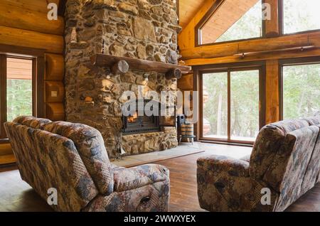 Divani imbottiti con pietra di campo illuminata e caminetto in roccia porosa nel soggiorno all'interno di una capanna in legno di cedro rosso artigianale, Quebec, Canada Foto Stock