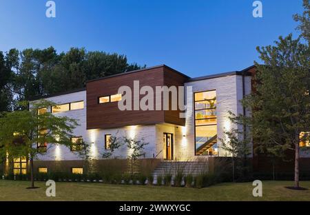 Pietra beige illuminata con pareti in legno di cedro marrone che si affacciano su una casa moderna in stile cubista con giardino panoramico al crepuscolo in estate, Quebec, Canada Foto Stock