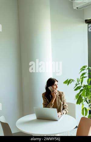 Una donna siede a un tavolo bianco in un ufficio illuminato dal sole, perso nel pensiero, con un portatile e una pianta verde accanto a lei. Foto Stock