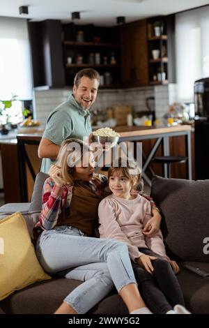 Una famiglia di tre persone si trova comodamente adagiata su un divano, con i volti che riflettono l'emozione e l'attenzione mentre condividono una ciotola di popcorn durante una sosp Foto Stock