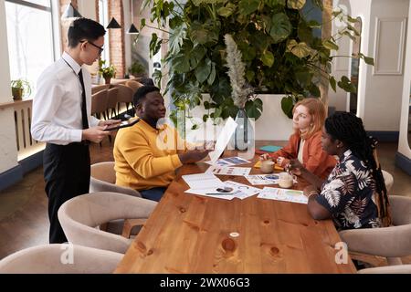 Giovane uomo di colore che ordina cibo durante una riunione di lavoro con diversi colleghi in un ristorante moderno, cameriere asiatico con tablet digitale che prende l'ordine Foto Stock