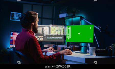 Editor video che utilizza un software di editing su monitor con schermo verde per aggiornare le riprese, commissionato dai team di produzione che esternalizzano le attività. Un operatore video freelance che completa il progetto su un PC mockup Foto Stock