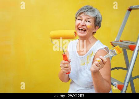 Senior felice seduto su una scala a cavalletto con un pennello e un rotolo di vernice vicino a una parete gialla Foto Stock
