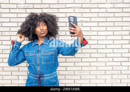 Questa vivace immagine cattura una giovane donna afroamericana con un abito di tendenza in denim che scatta un selfie. Il suo allegro pupazzo e la voluminosa acconciatura afro si distinguono contro la parete in mattoni bianchi testurizzati, incarnando un momento di stile urbano ed espressione di sé. Elegante donna afroamericana che porta Selfie contro White Brick Wall. Foto di alta qualità Foto Stock