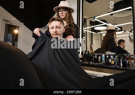 Un giovane con la barba in un mantello nero siede su una sedia in un barbiere. Il cliente sta aspettando un taglio di capelli e la rasatura della barba. Foto Stock