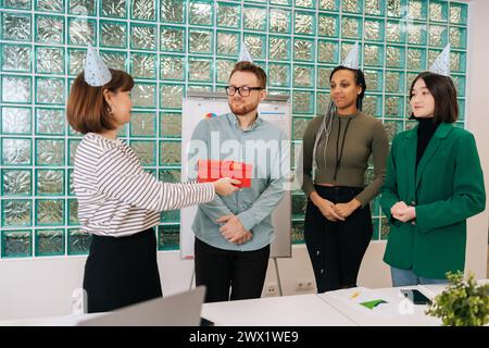 Felice gruppo multietnico di dipendenti della startup che festeggiano il compleanno regalando una confezione regalo a un giovane e affascinante collega. Foto Stock
