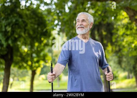 Ritratto di un vecchio uomo dai capelli grigi in piedi in un parco con pali da trekking, facendo nordic walking. Non guarda con un sorriso. Foto Stock