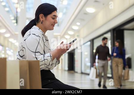 Vista laterale del ritratto di una donna mediorientale che utilizza lo smartphone mentre è seduta sulla panchina nel centro commerciale, copia spazio Foto Stock