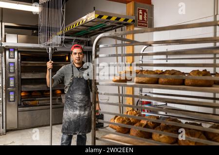 Il maestro latino baker prende i pani fuori dal forno e li mette al fresco, posa di ritratti e guarda la macchina fotografica Foto Stock