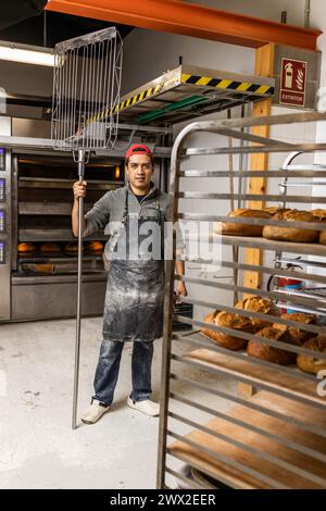 Il maestro latino baker prende i pani fuori dal forno e li mette al fresco, posa di ritratti e guarda la macchina fotografica Foto Stock