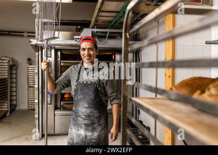 Il maestro latino baker prende i pani fuori dal forno e li mette al fresco, posa di ritratti e guarda la macchina fotografica Foto Stock