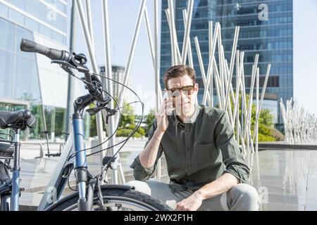 il giovane uomo d'affari sorridente guida una bicicletta elettrica mentre si dirige verso la torre dove si trova il suo ufficio, conversando con il tuo smartphone Foto Stock