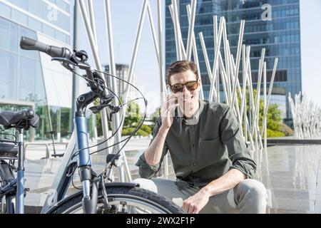 il giovane uomo d'affari sorridente guida una bicicletta elettrica mentre si dirige verso la torre dove si trova il suo ufficio, conversando con il tuo smartphone Foto Stock