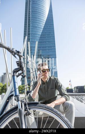 il giovane uomo d'affari sorridente guida una bicicletta elettrica mentre si dirige verso la torre dove si trova il suo ufficio, conversando con il tuo smartphone Foto Stock