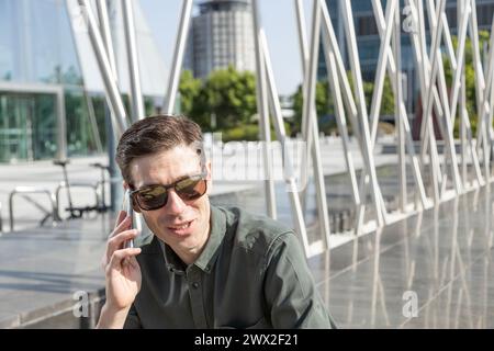 il giovane uomo d'affari sorridente guida una bicicletta elettrica mentre si dirige verso la torre dove si trova il suo ufficio, conversando con il tuo smartphone Foto Stock