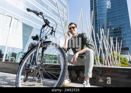 il giovane uomo d'affari sorridente guida una bicicletta elettrica mentre si dirige verso la torre dove si trova il suo ufficio, conversando con il tuo smartphone Foto Stock