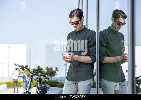 il giovane uomo d'affari sorridente guida una bicicletta elettrica mentre si dirige verso la torre dove si trova il suo ufficio, controllando i messaggi sul suo smartphone Foto Stock