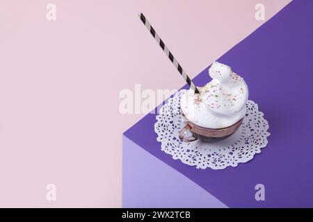 Un cubo viola realizzato con carta colorata per creare un'illusione ottica 3D. In cima c'è una tazza di caffè rosa vintage con panna montata e cospargi su una W Foto Stock