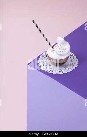 Un cubo viola realizzato con carta colorata per creare un'illusione ottica 3D. In cima c'è una tazza di caffè rosa vintage con panna montata e cospargi su una W Foto Stock