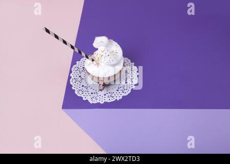 Un cubo viola realizzato con carta colorata per creare un'illusione ottica 3D. In cima c'è una tazza di caffè rosa vintage con panna montata e cospargi su un wh Foto Stock