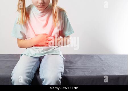 Una bambina di sette anni si siede e tiene la pancia rossa con le mani. Concetto di dolore addominale nei bambini. Infezione da enterovirus e poisonina Foto Stock