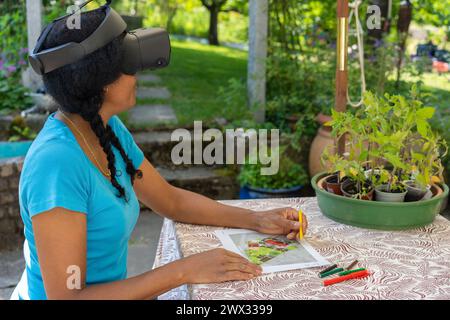 Una donna di corsa mista con occhiali VR progetta un giardino con matite colorate a un tavolo. Foto Stock