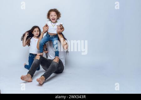 Giovane papà latino con i capelli lunghi e gli occhiali, seduto sul pavimento a giocare con le sue due figlie con i capelli ricci, indossando una t-shirt bianca e jean Pan Foto Stock