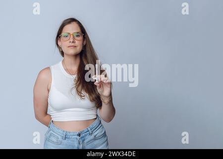 giovane donna latina medico estetista, sorridente guardando la macchina fotografica, indossando occhiali e vestiti bianchi, in piedi in posa con la siringa in mano, studio sh Foto Stock